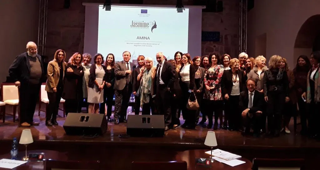 AMINA participants and partners gathered on stage at a programme event, the AMINA title on the screen behind them
